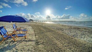 Ein Strand mit blauen Liegestühlen und einem blauen Sonnenschirm, im Hintergrund ein Steg und die Sonne scheint.