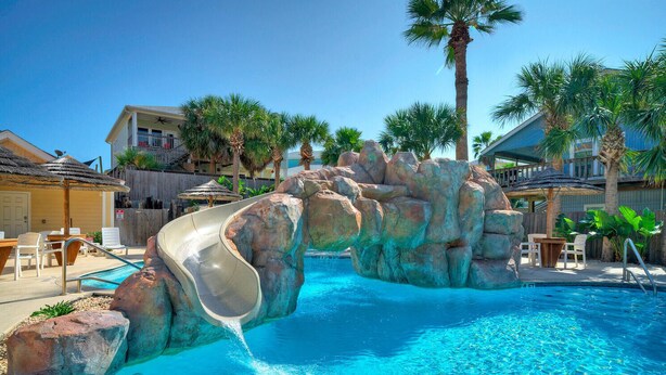 Une piscine avec un toboggan, des palmiers et une terrasse en bois avec des chaises.