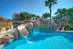 A pool with a water slide, palm trees, and a wooden deck with chairs.