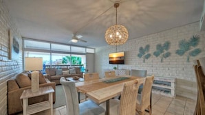 Un salón amplio con una mesa de comedor, sillas de mimbre, un ventilador de techo y un ventanal que ofrece vistas al exterior.