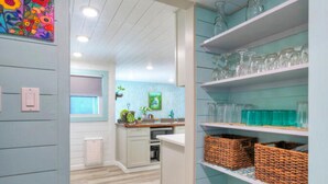 A kitchen with light blue walls, wooden floors, and shelves with glassware and baskets. - Big Yard w/ Priv Pool, Firepit, Patio | Near the Beach | Reel Time by AvantStay (Port Aransas)