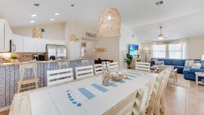 A spacious kitchen with white cabinets, a central island with seating, and a dining area with a striped tablecloth. - Salty Peach by AvantStay | Private Pool, New Decor (Port Aransas)