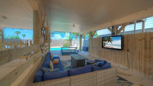 An outdoor patio with a pool, wooden deck, and a TV mounted on the wall. - Salty Peach by AvantStay | Private Pool, New Decor (Port Aransas)