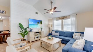 A living room with a blue sofa, wooden coffee table, and a television displaying a beach scene. - Salty Peach by AvantStay | Private Pool, New Decor (Port Aransas)