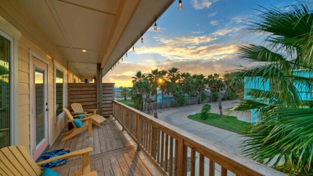 A wooden deck with chairs and a view of palm trees and a road. - Pool, Walk to Old Town + Eateries, Nightlife, Shopping | Rock N Reelby AvantStay (Port Aransas)