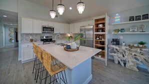 A modern kitchen with white cabinetry, a central island with bar stools, a microwave, a refrigerator, and pendant lighting. - Outdoor Lounge w/ Bar Swing Seats, Near Nature Preserve | Blutopia by AvantStay (Port Aransas)