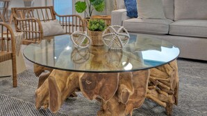 A round glass coffee table with a wooden base, surrounded by wicker chairs and a sofa in a modern living room. - Outdoor Lounge w/ Bar Swing Seats, Near Nature Preserve | Blutopia by AvantStay (Port Aransas)