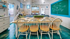 A dining area with a wooden table and white chairs, a chalkboard with a compass drawing, and a view of the beach through a window. - Stella Maris by AvantStay | Rooftop View + Pool (Port Aransas)