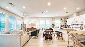 A spacious living area with a sofa, dining table, and chairs, a kitchen with white cabinets and a microwave, and large windows with blinds. - Newly Built, Pool, Walk to Old Town Eats+Marina | Shorely Goodness by AvantStay (Port Aransas)