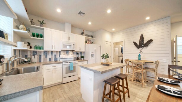 A modern kitchen with white cabinetry, a central island, wooden stools, and a dining table. - Newly Built, Pool, Walk to Old Town Eats+Marina | Shorely Goodness by AvantStay (Port Aransas)