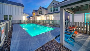 A swimming pool surrounded by a wooden fence, with a covered seating area and a building with multiple windows. - Newly Built, Pool, Walk to Old Town Eats+Marina | Shorely Goodness by AvantStay (Port Aransas)