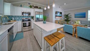 Cozinha moderna com armários brancos, eletrodomésticos de aço inoxidável e uma ilha central. Há uma área de jantar com um sofá azul e cadeiras de madeira.
