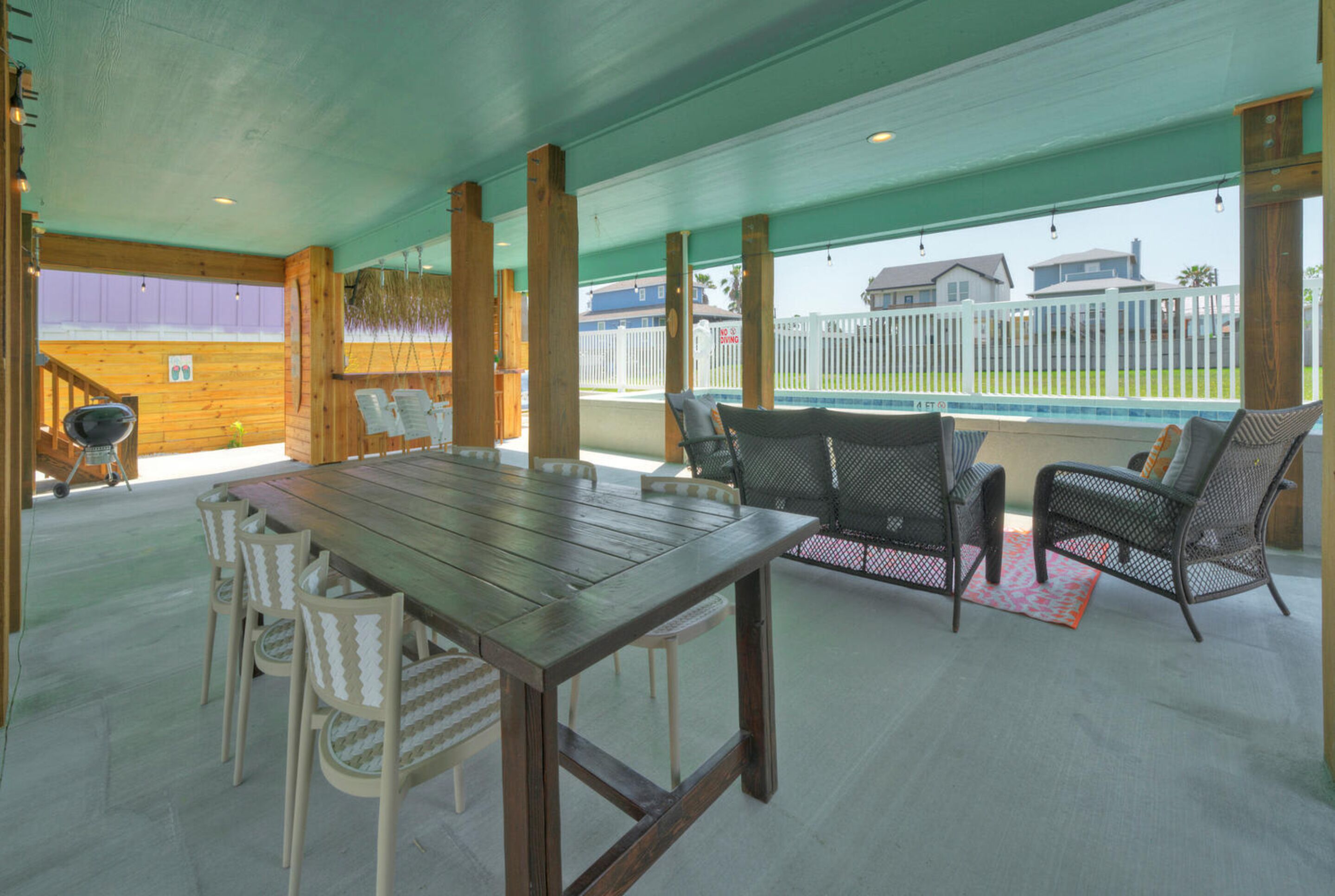Une terrasse extérieure avec une table et des chaises en bois, une piscine et une vue sur les maisons au loin.
