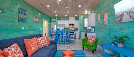 A room with a wooden ceiling, a ceiling fan, a blue sofa with orange cushions, a green armchair, and a kitchen area with white cabinets and blue chairs.