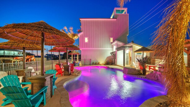 A swimming pool with a curved edge, surrounded by lounge chairs and umbrellas, and a building with a pink facade in the background.
