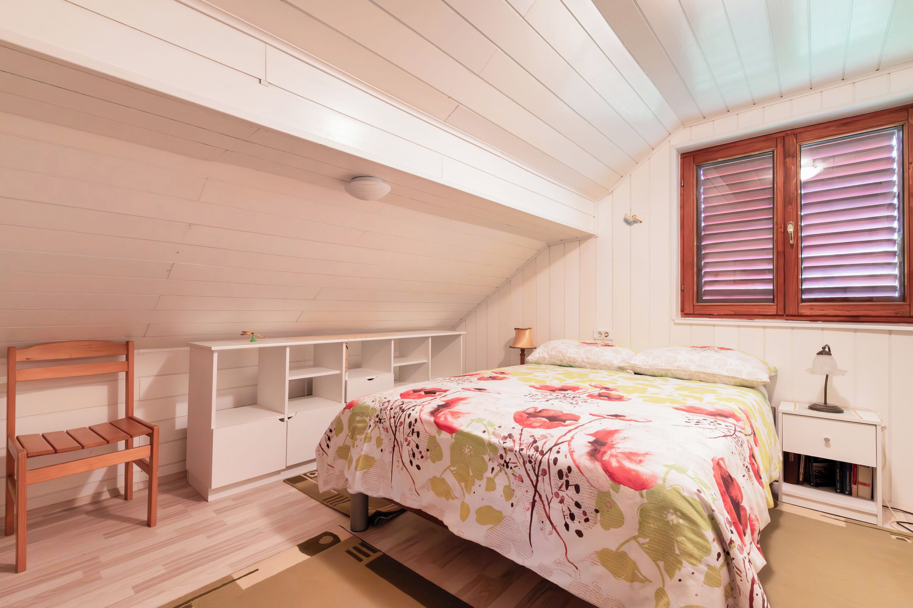 A bedroom with a bed, a wooden chair, a white shelving unit, and a window with blinds.