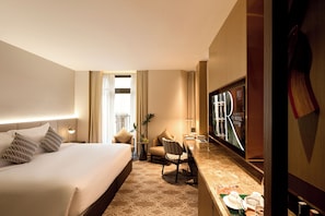 A hotel room with a large bed, a desk with a chair, a television, and a small table with a plant and a vase.