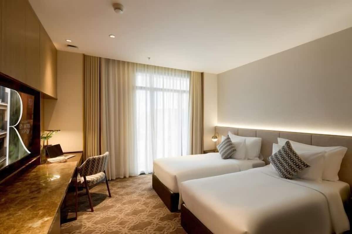 A hotel room with two beds, a desk with a chair, a TV, and a large window with curtains.
