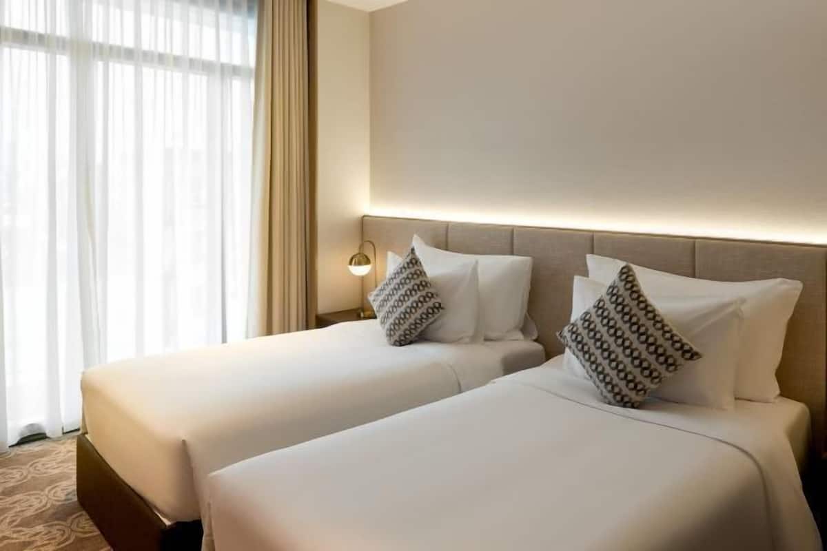A hotel room with two beds, a headboard, a nightstand, a lamp, and a window with curtains.