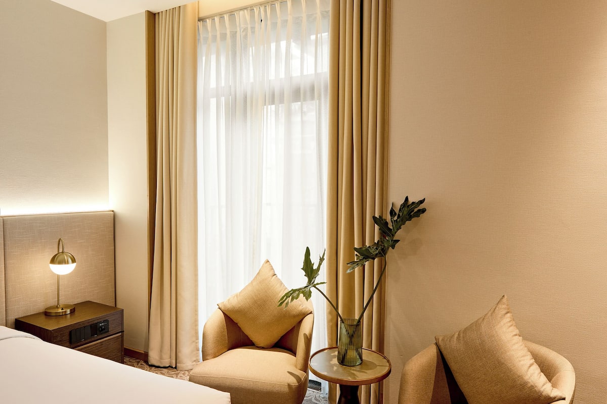 A hotel room with a bed, two armchairs, a small table with a vase, and a lamp.