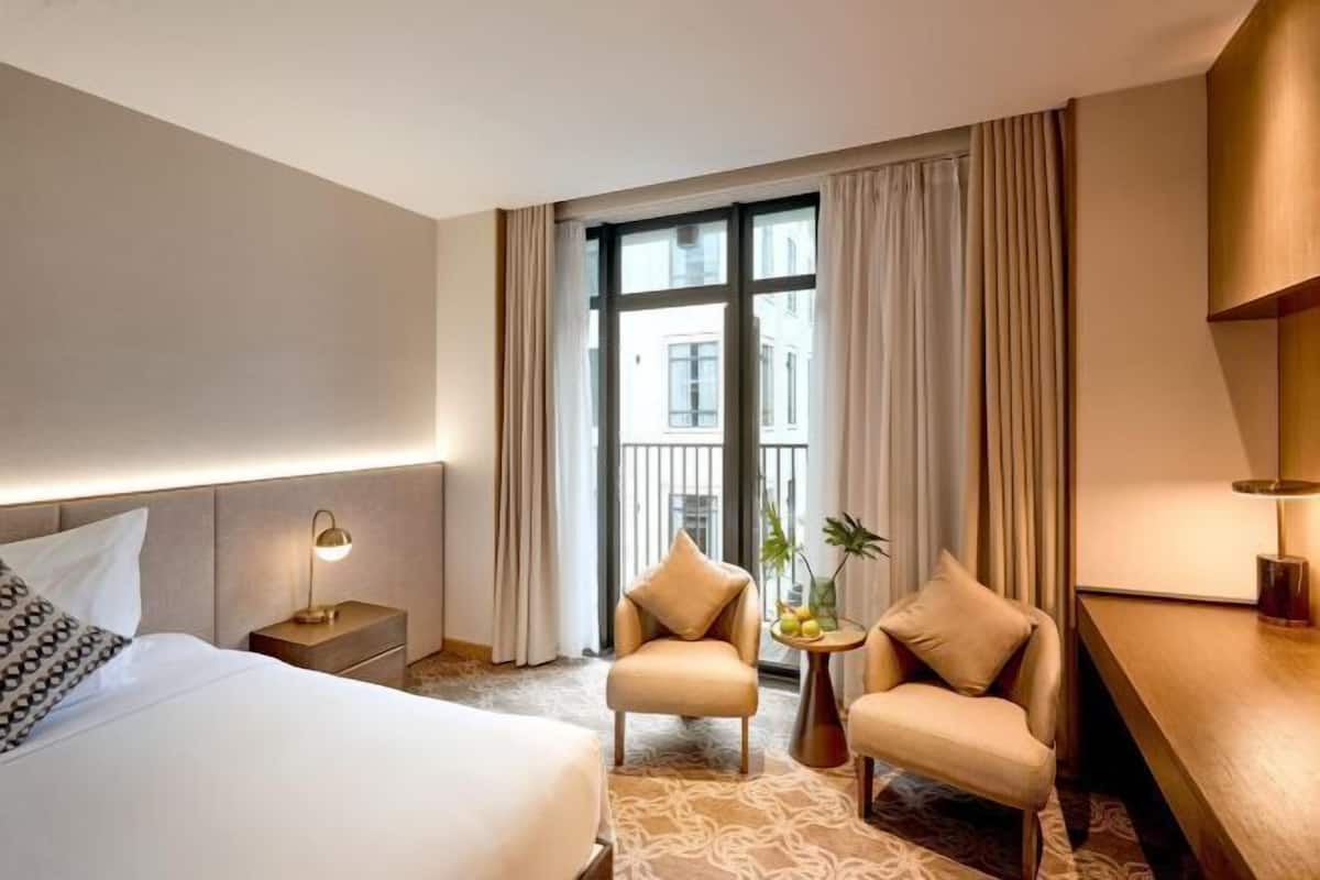 A hotel room with a bed, two armchairs, a small table with a plant, and a view of a building through large windows.