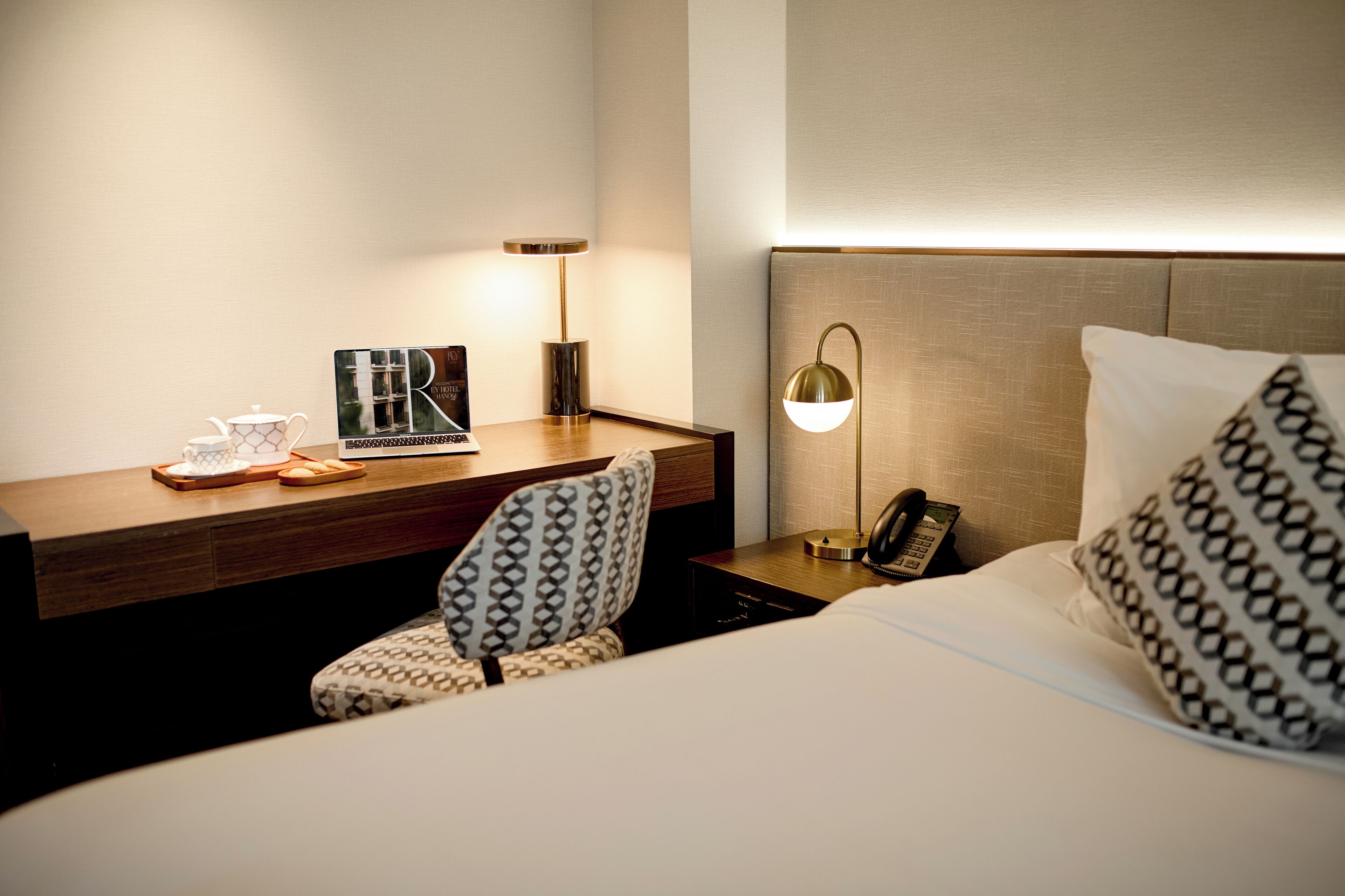 A hotel room with a neatly made bed, a bedside table with a lamp, a chair, and a small table with a teapot and cups.