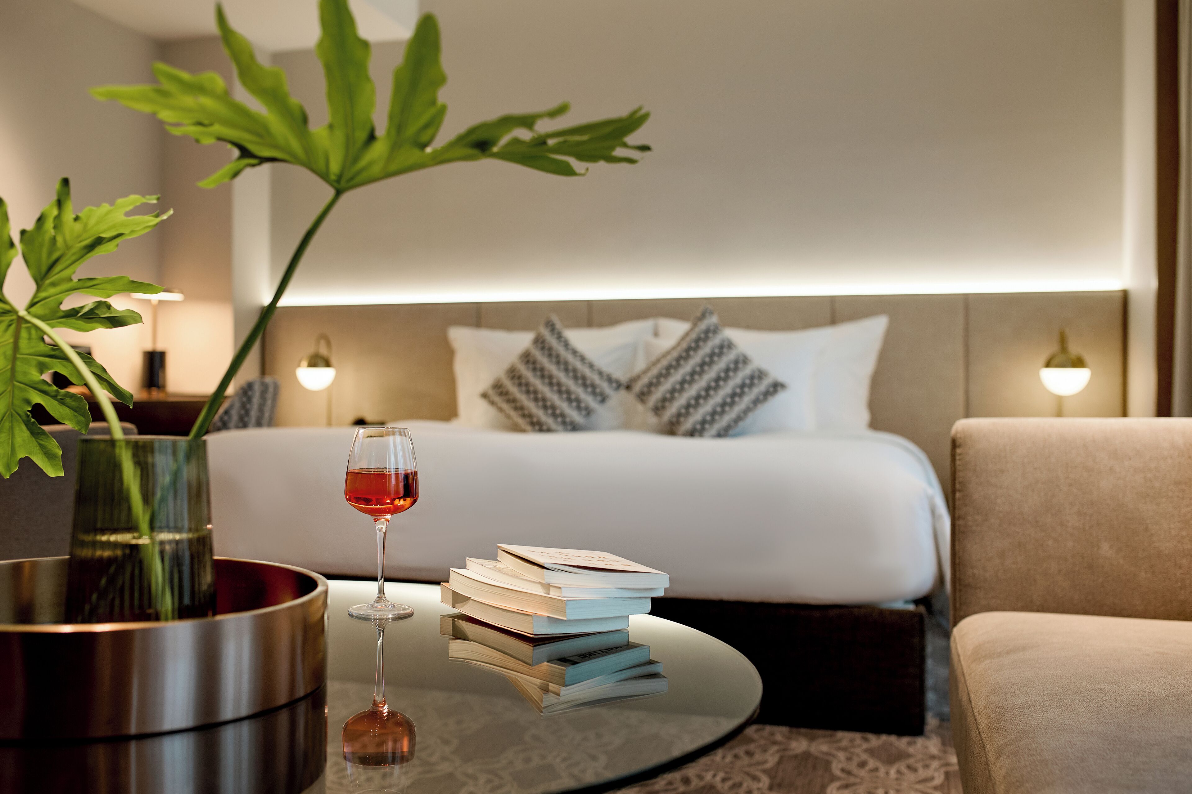 Ein Hotelzimmer mit einem Bett, Nachttischlampen, einem Couchtisch mit Büchern und einem Glas Rotwein sowie einer Topfpflanze.