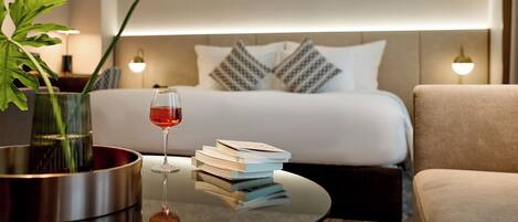 Ein Hotelzimmer mit einem Bett, Nachttischlampen, einem Couchtisch mit Büchern und einem Glas Rotwein sowie einer Topfpflanze.