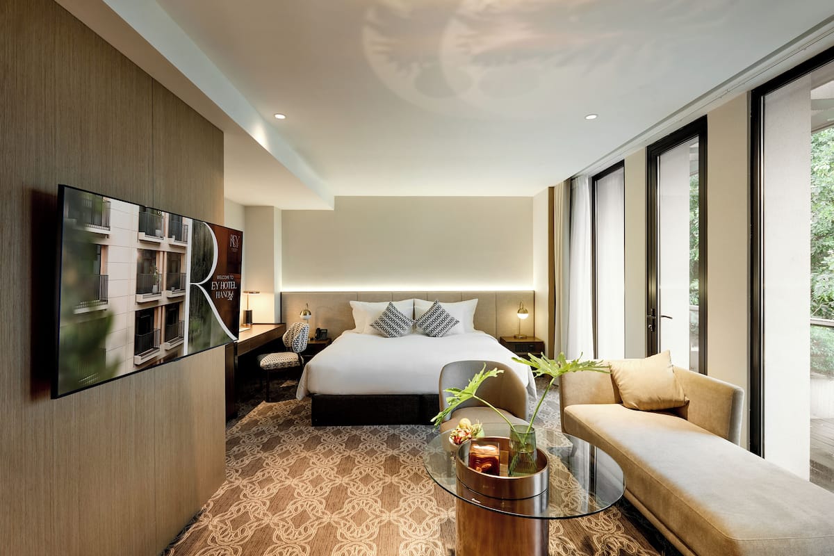 A modern hotel room with a large bed, a sofa, a TV mounted on the wall, and a large window offering a view of the outdoors.