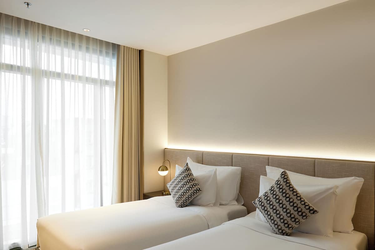 A hotel room with two beds, a large window with sheer curtains, a patterned carpet, and a wall-mounted lamp.