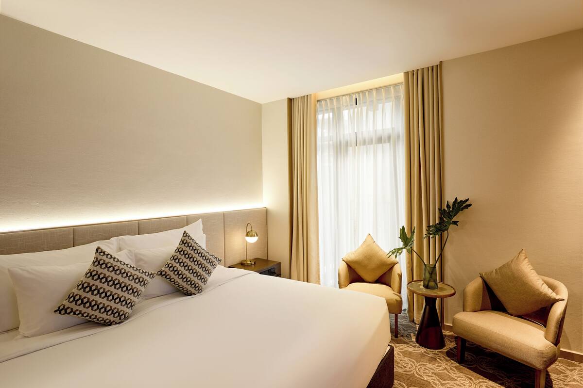 A hotel room with a large bed, two bedside tables, a lamp, a chair, and a small table with a plant.
