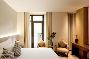 A hotel room with a bed, two armchairs, a small table, a plant, and a desk with a lamp.