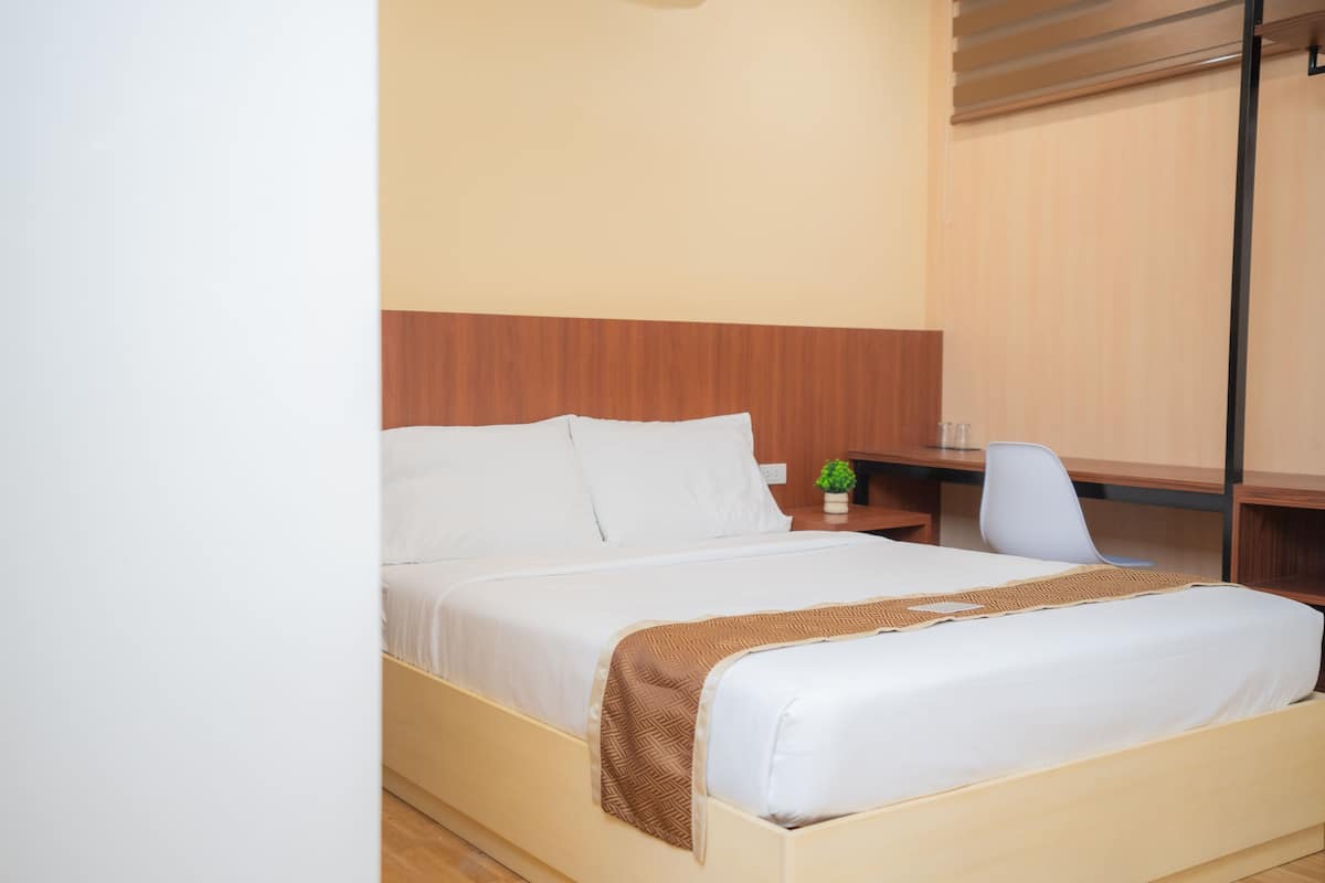 A neatly made bed with white linens and a brown throw blanket, a small bedside table with a green plant, and a chair next to a wooden desk.