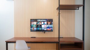 A modern room with a wooden desk, a white chair, and a wall-mounted TV displaying various shows. - Sundaze Dormitel (Naga)