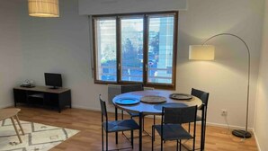 Un coin repas comprenant une table ronde, quatre chaises, un téléviseur posé sur un bureau et une fenêtre avec des stores.