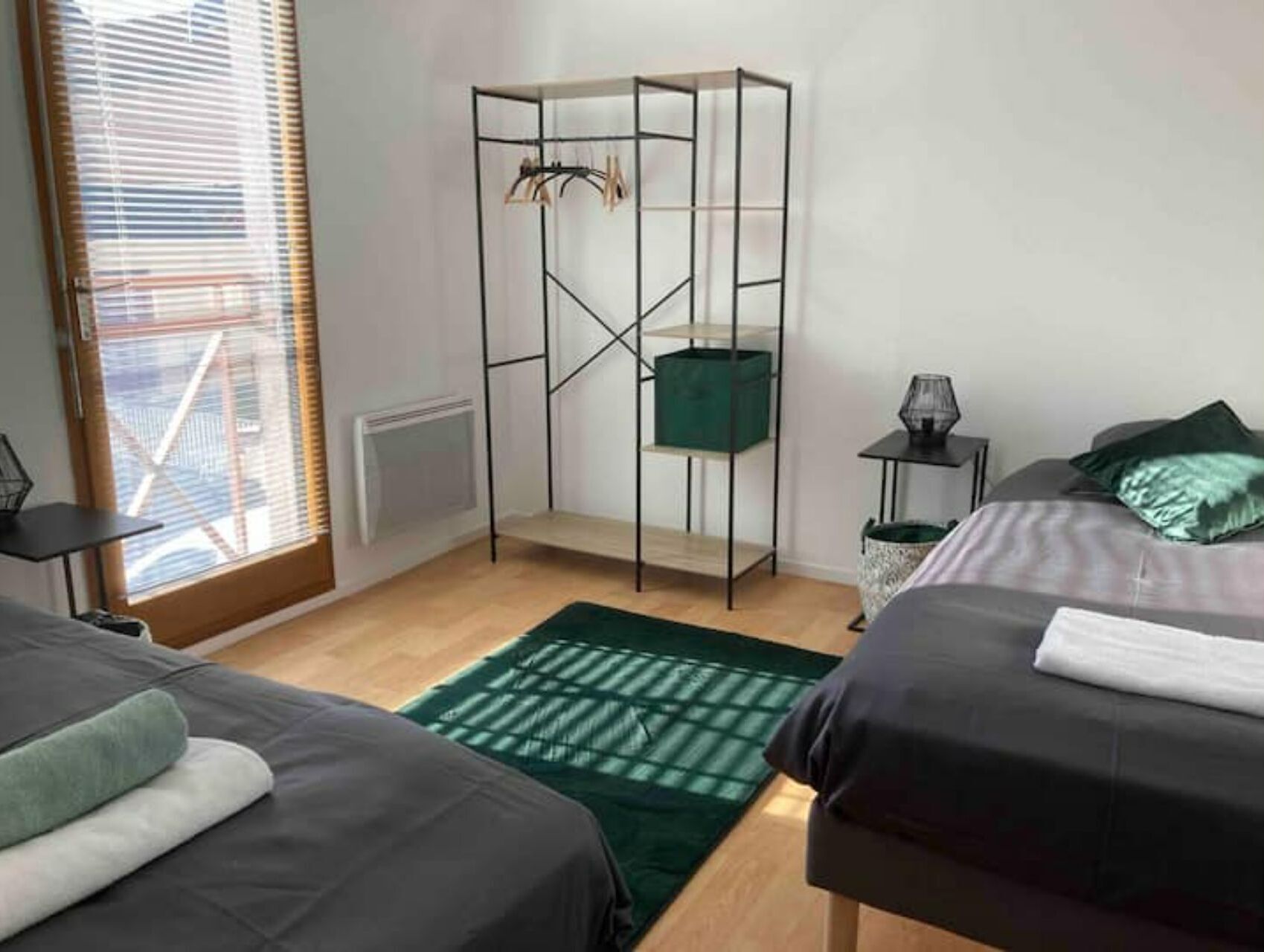 Een slaapkamer met twee bedden, een groen vloerkleed, een radiator, een raam met jaloezieën, een metalen plankkast en een kleine tafel met een lamp.