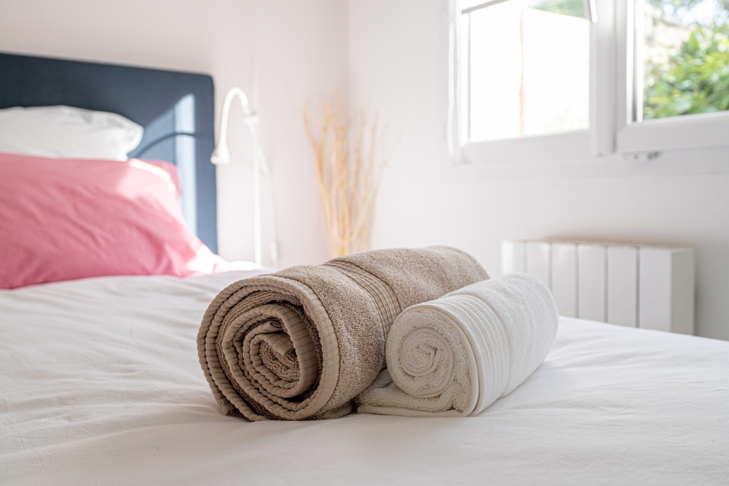 Una cama bien hecha con toallas enrolladas y una almohada rosa, un radiador blanco y una ventana con vista a follaje.