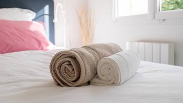 Una cama bien hecha con toallas enrolladas y una almohada rosa, un radiador blanco y una ventana con vista a follaje.