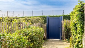 Une entrée fermée par un portail, avec une porte bleue, entourée de verdure et d’une clôture en bois.