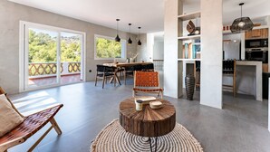 Un vaste espace de vie comprenant une table à manger, des chaises, une petite table basse ronde et une grande fenêtre donnant sur un paysage verdoyant.