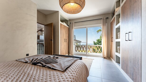 Une chambre dotée d’un grand lit, d’un luminaire au plafond, d’une porte coulissante donnant sur un balcon et d’armoires encastrées en bois.