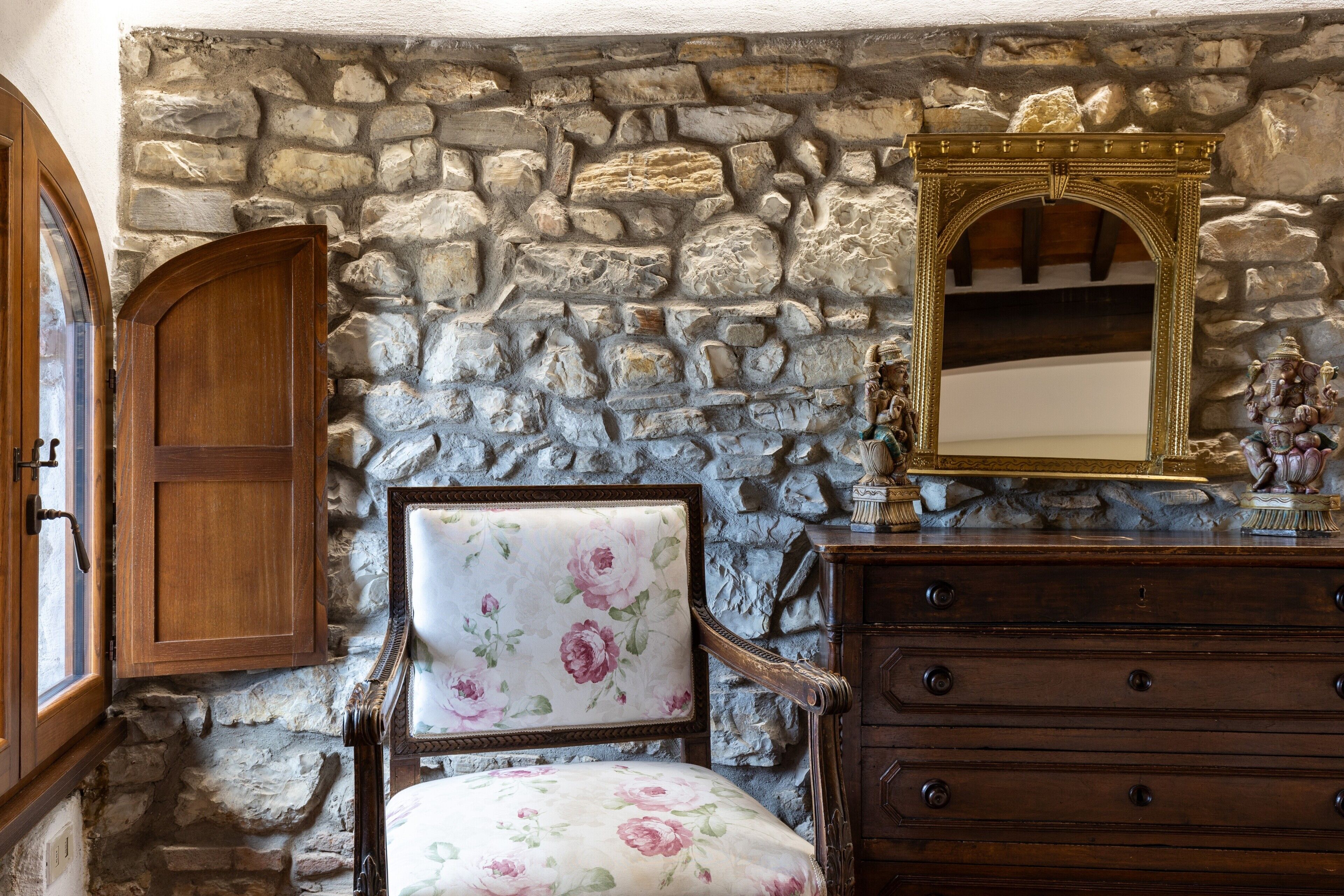 A room with a stone wall, a wooden cabinet, a floral upholstered chair, and a wooden dresser with drawers.