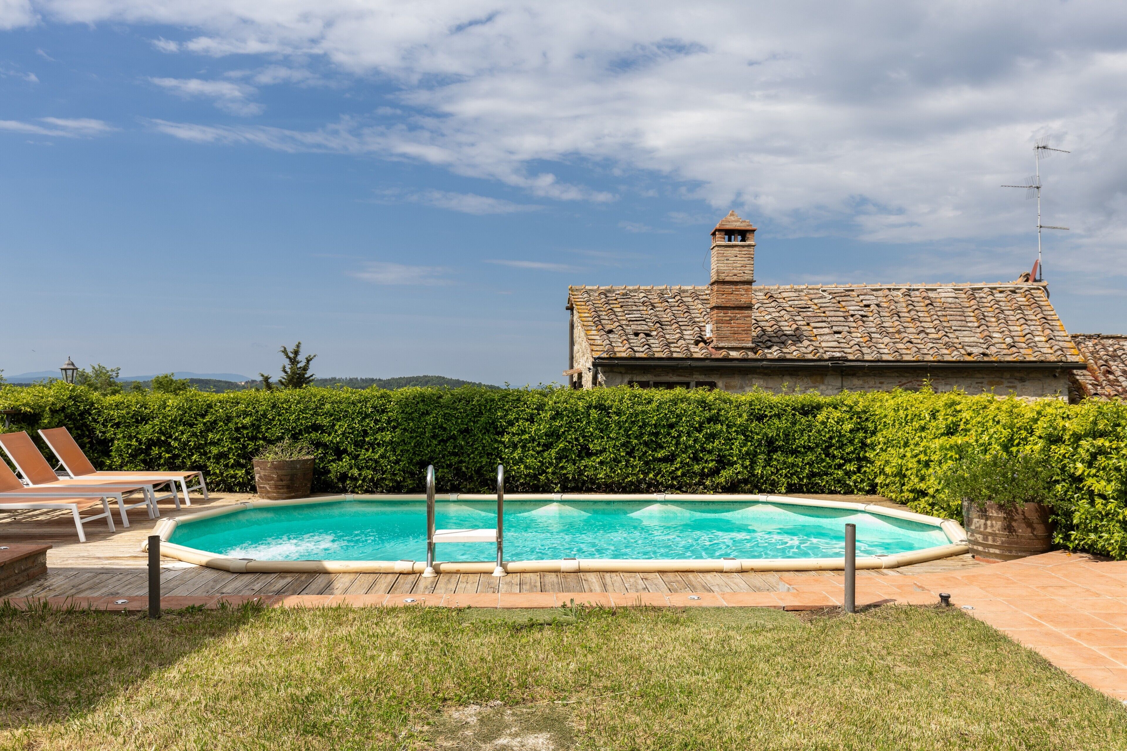 A swimming pool with a wooden deck, a brick house with a chimney, and a clear sky.