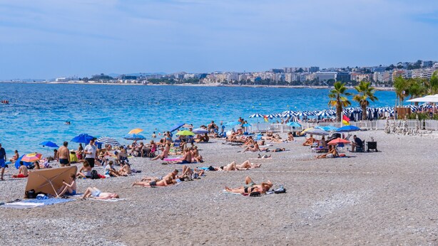 A beach with people sunbathing and swimming, umbrellas, and a cityscape in the background.