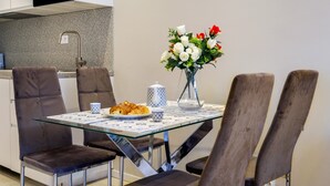 A dining area with a glass table, four chairs, a vase with flowers, a croissant, and a teapot.