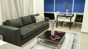 A modern living room with a dark green sofa, a glass coffee table with books, a vase with flowers, and a dining area with a blue wall and white chairs. - Blue (Beirut)