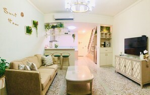 Una sala de estar con sofá, mesa de centro y televisor. Hay plantas en las paredes y una cocina al fondo.
