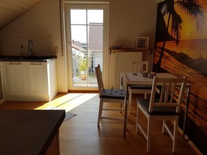 Une cuisine avec des meubles blancs, un coin repas avec une table et des chaises, une vue sur l’extérieur grâce à une porte vitrée, et un tableau représentant un coucher de soleil accroché au mur.