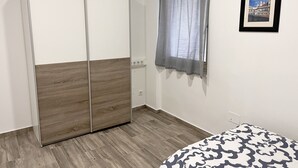 A room with a wardrobe, a window with curtains, a bed with a patterned bedspread, and a framed picture on the wall.