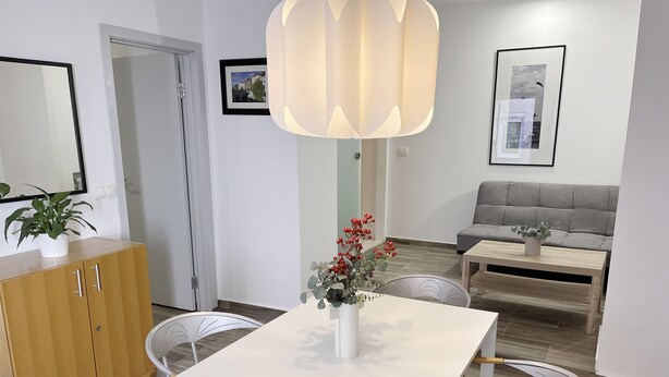 A modern living room with a white dining table, two chairs, a grey sofa, and a wooden cabinet.